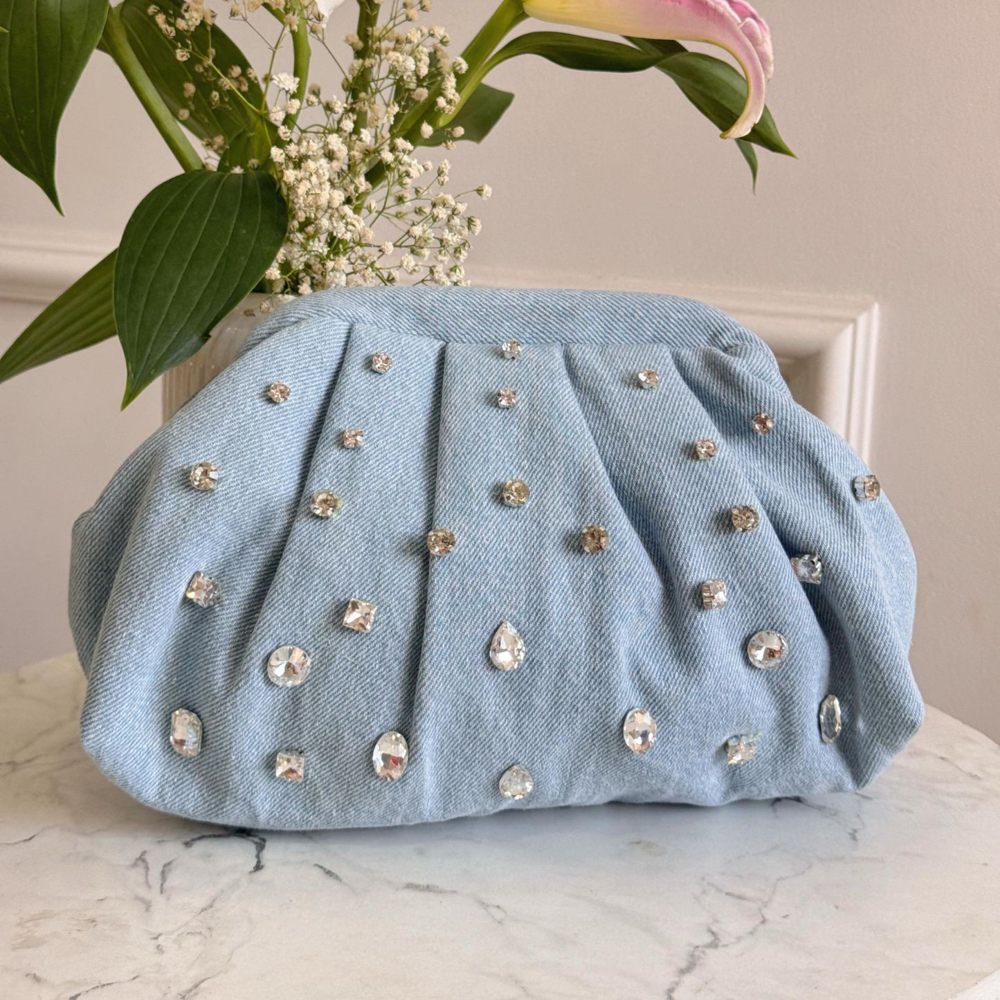 Light blue clutch bag with gemstone embellishments on a marble surface with flowers in the background