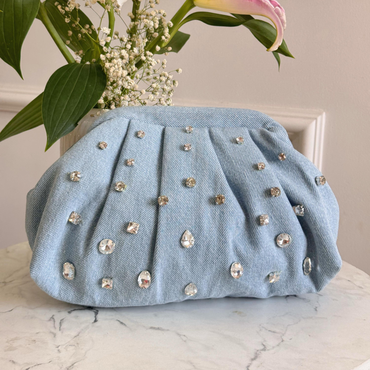 Light blue clutch bag with gemstone embellishments on a marble surface with flowers in the background